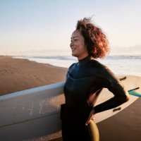 side view portrait of a woman standing in the surf with a surfboard under her arm. - travel stock pictures, royalty-free photos & images