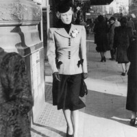 Smartly dressed woman walks down a London street. Her tailored suit has squared shoulders and the contrasting buttons and trim on her jacket match...