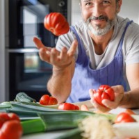 smiling 50 year old man with a beard in front of a table full of fresh vegetables tossing a tomato. healthy food concept. - food stock pictures, royalty-free photos & images