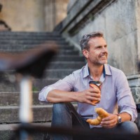smiling man sitting on stairs with croissant and takeaway coffee in the city - junk food stock pictures, royalty-free photos & images