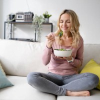 smiling woman with bowl of salad sitting on sofa - food stock pictures, royalty-free photos & images