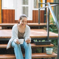 smiling woman with curly hair in casual clothes with cup of morning coffee using mobile phone sitting at backyard of house - garden decoration photos et images de collection