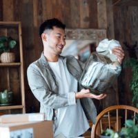 smiling young asian man looking happy as he unboxing the delivered package from online purchases at home, take out a glass vase. online shopping, enjoyable customer shopping experience - home decoration stock pictures, royalt