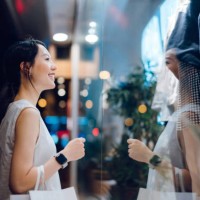 smiling young asian woman carrying a paper shopping bag, standing outside a boutique looking at shop window in the evening in city - fashion stock pictures, royalty-free photos & images