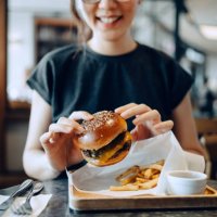 smiling young asian woman enjoying freshly made delicious burger with fries and a glass of iced coffee in a cafe - junk food stock pictures, royalty-free photos & images
