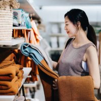 smiling young asian woman shopping for home decor and necessities in a homeware store, choosing for a blanket on a shelf - home decoration stock pictures, royalty-free photos & images