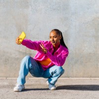 smiling young dancer in squatting position taking selfie through mobile phone in front of wall - fashion stock pictures, royalty-free photos & images