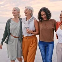 strand, freunde und urlaub mit einer älteren frauengruppe, die auf dem sand am meer oder ozean spazieren geht. natur, wasser und freundschaft mit vielfalt und reifen weibchen, die bei sonnenuntergang in ihrem ruhestand lachen