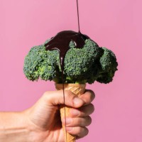 studio shot of man's hand holding ice cream cone with broccoli on top and pouring chocolate sauce, against pink background - food stock pictures, royalty-free photos & images