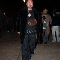 Takashi Murakami wears glasses, a black fluffy coat, a necklace depicting a cartoon character, a black pullover with silver shiny Louis Vuitton...