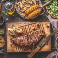 tasty grilled bbq meat ribs on wooden cutting board and grill grate with knife and fresh garlic herb butter bread served on rustic table with grilled corn cobs with bbq sauce , salad leaves and other tasty ingredients - food 