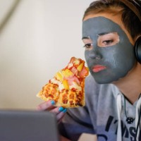 teenage girl wearing headphones and watching her laptop while holding a slice of pizza. - junk food stock pictures, royalty-free photos & images