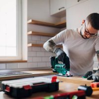 the bearded carpenter using electric jigsaw for cutting a wooden plank in his new kitchen. - home decoration stock pictures, royalty-free photos & images