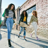three young women with long curly hair walking along pavement, one carrying skateboard. - fashion stock pictures, royalty-free photos & images