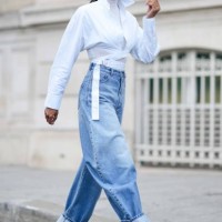 Tina Kunakey wears gold earrings, a white turtleneck long sleeves t-shirt, a white laces waist cropped waist shirt, high waist blue faded denim puffy...
