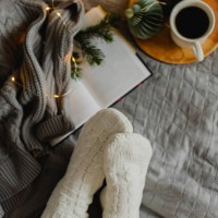 top view of female feet in cozy white socks on bed with cup of coffee, christmas lights and open book. christmas, relaxation and hygge concept. - home decoration stock pictures, royalty-free photos & images