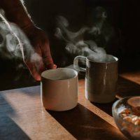 two cups of coffee on a wooden table with a bowl of breakfast cereal. someone lifts one of the mugs out of frame. - coffee stock pictures, royalty-free photos & images
