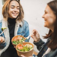 two females friends eating take away vegetarian bowls outdoors in town. - junk food stock pictures, royalty-free photos & images