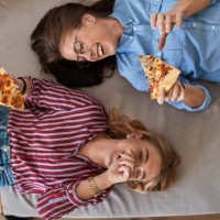 two laughing young women lying down eating pizza together - food stock pictures, royalty-free photos & images