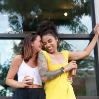 two millennial women, one caucasian with shoulder length hair and the other mixed race with curly hair, laugh while they hug each other outdoor near a building, wearing casual summer clothing. - fashion stock pictures, royalt