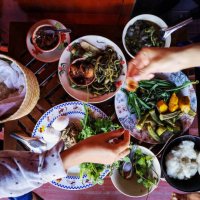 two thai women traditionally eating a selection of freshly cooked northern thai food of vegetables, soups and curries served in dishes on a wooden table, along with sticky rice, which is a food staple in northern and northeas