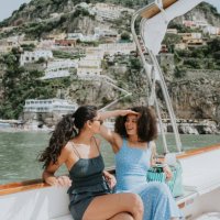 two woman enjoy a journey on a boat, with positano, italy, in the background. one woman wears sunglasses, the other shields her eyes from the bright light with her hands. - travel stock-fotos und bilder