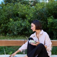 vertical photo of a businesswoman sitting on an outdoor bench, eating a healthy salad during her lunch break while working on a laptop computer - junk food stock pictures, royalty-free photos & images