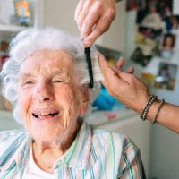 weitwinkelaufnahme einer fröhlichen, niedlichen älteren kaukasischen frau zu hause, die sich von einem professionellen stylisten für häusliche pflegekräfte die haare kämmen lässt - hairstyle stock-fotos und bilder