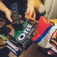 With remote control in one hand and nacho chip in the other, a teenager eats the food she likes in front of the TV.