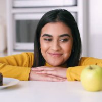 woman choosing between a cake or apple in the kitchen to eat as a day time snack at home. indian girl thinking about the healthy, nutrition and organic fruit or the unhealthy chocolate dessert. - junk food stock pictures, roy