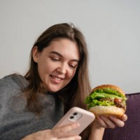 woman eating a big burger with meat, tomatoes and lettuce sitting on the couch at home, looking at the phone and smiling, concept of fast food - junk food stock pictures, royalty-free photos & images