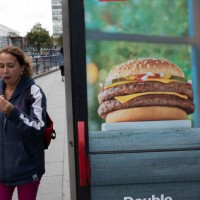 Woman eating a burger while passing an advertisement for burgers at Elephant and Castle in London, UK. The area is now subject to a master-planned...