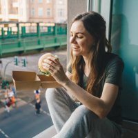 woman eating take out burger at home in front of the open window - junk food stock pictures, royalty-free photos & images