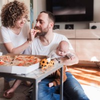 woman feeding pizza to her husband who is holding a sleeping baby - junk food stock pictures, royalty-free photos & images