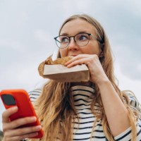 woman holds a smartphone in her hands and eats a bun with appetite on a city street - junk food stock pictures, royalty-free photos & images