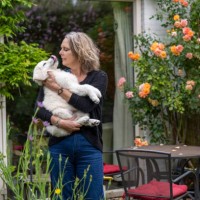 woman standing in a garden holding a white dog with blooming roses and outdoor furniture in the background - garden decoration photos et images de collection