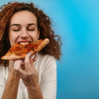 woman standing in front of blue background and eating pizza - junk food stock pictures, royalty-free photos & images