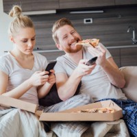 woman text messaging and man eating pizza and holding remote control - junk food stock pictures, royalty-free photos & images
