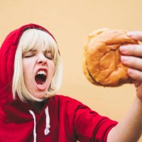woman with eyes closed holding hamburger in front of peach wall - junk food stock-fotos und bilder