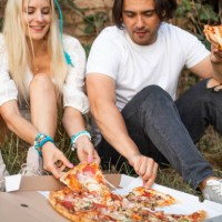 wonderful couple sitting on ground outdoors and taking pieces of fresh pizza from box. - junk food stock pictures, royalty-free photos & images