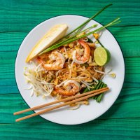 world famous freshly stir-fried cooked, thai recipe of prawn pad thai noodles on a round traditional enameled metal dish with chopsticks laid on the side of the dish, set on an abstract weathered turquoise colored wood panel 