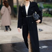 Xenia Adonts wears white mini dress, black long coat, black Chanel belt, black Chanel bag, beige over the knees Chanel boots, outside Chanel, during...