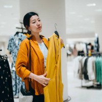 young asian woman shopping in a clothing store, looking at a yellow dress in mirror. sale season. seasonal shopping - fashion stock pictures, royalty-free photos & images