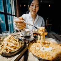 young asian woman sitting at a table by the window enjoying the warmth of sunlight and having meal joyfully in a restaurant - food stock pictures, royalty-free photos & images