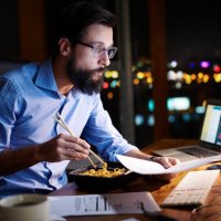 young businessman looking at computer and eating takeaway at office desk at night - junk food stock pictures, royalty-free photos & images