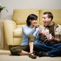 young couple sitting cross-legged in front of sofa eating takeaway food, woman feeding man with chopsticks, at home - junk food stock pictures, royalty-free photos & images