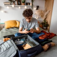 young man sitting on his bed, using smart phone and packing suitcase - travel stock pictures, royalty-free photos & images