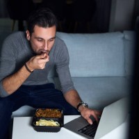 young man sitting on sofa in evening eating takeaway and using laptop - junk food stock pictures, royalty-free photos & images