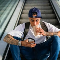 young man with backpack sitting on escalator looking at cell phone while drinking coffee to go - junk food stock pictures, royalty-free photos & images
