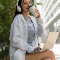 young smiling woman in headphones working on laptop and drinking takeaway coffee while sitting outdoors - junk food stock pictures, royalty-free photos & images